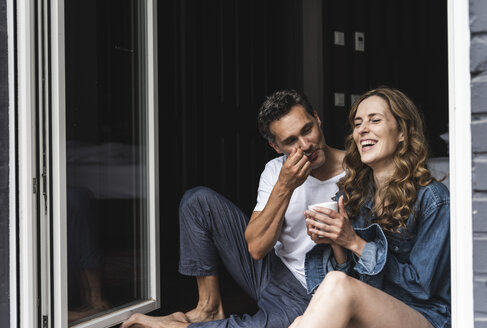 Happy couple in nightwear at home sitting at French window - UUF14340