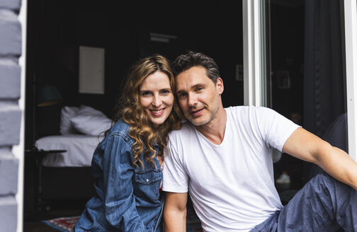 Portrait of smiling couple in nightwear at home sitting at French window - UUF14342