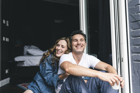 Happy couple in nightwear at home sitting at French window - UUF14345