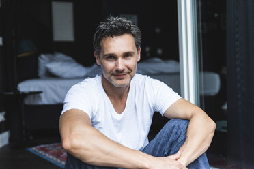 Portrait of confident man in pyjama at home sitting at French window - UUF14348