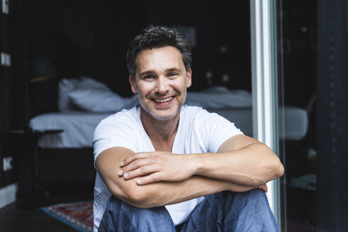 Portrait of smiling man in pyjama at home sitting at French window - UUF14349