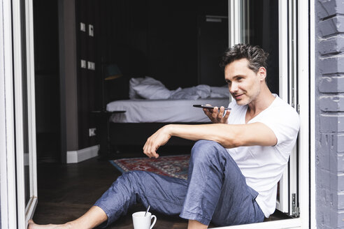 Mann im Pyjama zu Hause am französischen Fenster sitzend und mit Smartphone - UUF14351