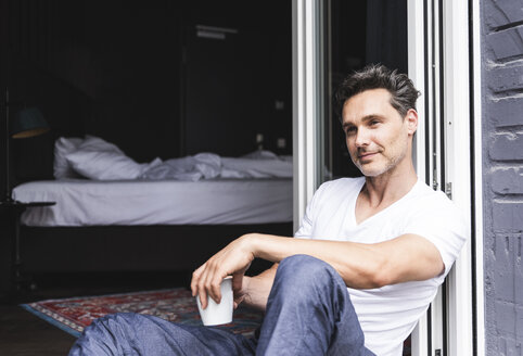 Smiling man in pyjama at home with cup of coffee sitting at French window - UUF14352