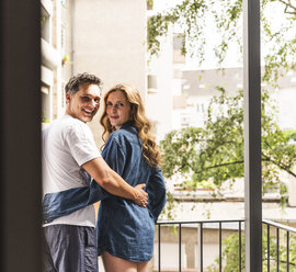 Glückliches Paar in Nachtwäsche auf dem Balkon stehend und sich umarmend - UUF14367