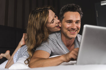 Glückliches Paar auf dem Bett liegend zu Hause mit Laptop - UUF14375