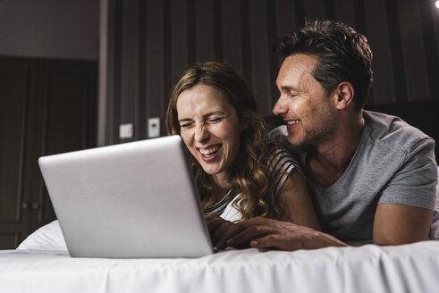 Glückliches Paar, das zu Hause auf dem Bett liegt und Spaß mit dem Laptop hat - UUF14377