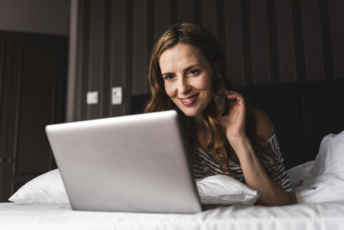 Lächelnde Frau, die zu Hause auf dem Bett liegt und auf ihren Laptop schaut - UUF14378