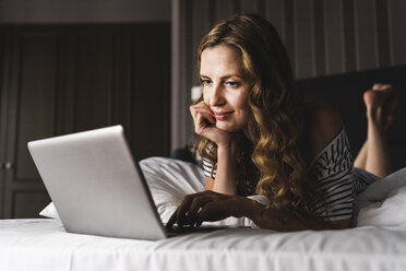 Smiling woman lying on bed at home looking at laptop - UUF14379