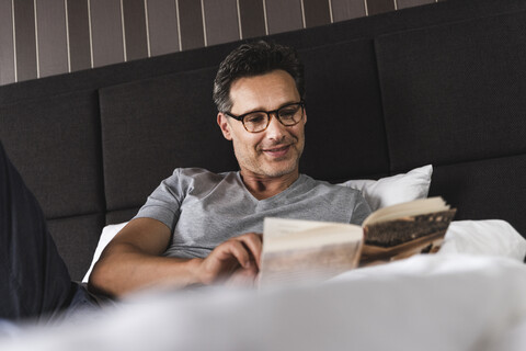 Lächelnder Mann, der zu Hause im Bett liegt und ein Buch liest, lizenzfreies Stockfoto