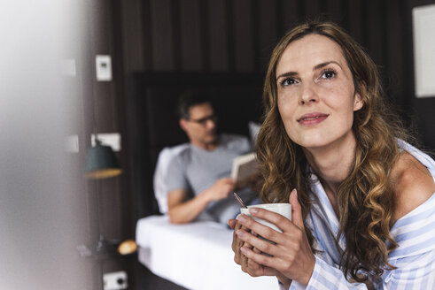 Lächelnde Frau mit einer Tasse Kaffee im Schlafzimmer mit einem Mann im Hintergrund - UUF14386