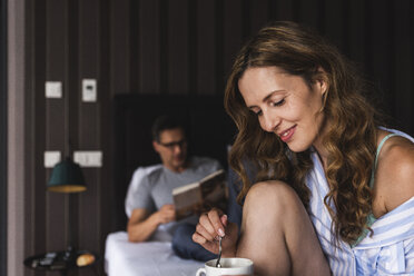 Lächelnde Frau mit einer Tasse Kaffee im Schlafzimmer mit einem Mann im Hintergrund - UUF14387