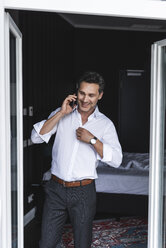 Lächelnder Geschäftsmann am Handy im Schlafzimmer zu Hause beim Anziehen - UUF14391