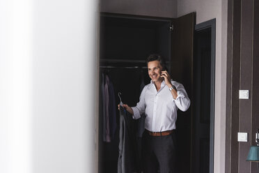 Smiling businessman on cell phone at home getting dressed - UUF14393