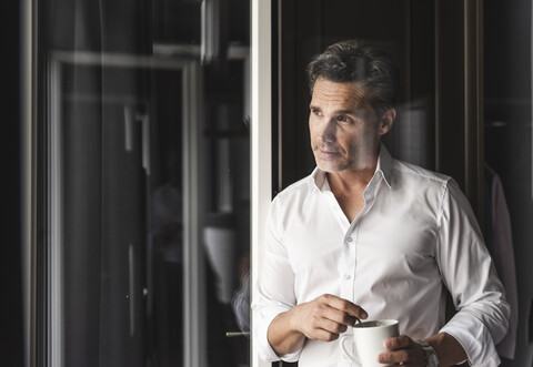 Geschäftsmann mit einer Tasse Kaffee, der zu Hause aus dem Fenster schaut, lizenzfreies Stockfoto