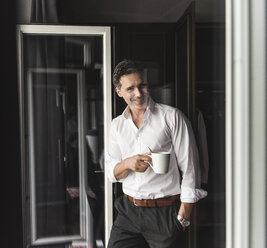Smiling businessman with cup of coffee standing at the window at home - UUF14402