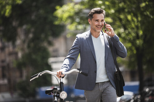 Lächelnder Mann mit Handy beim Schieben eines Fahrrads in der Stadt - UUF14417