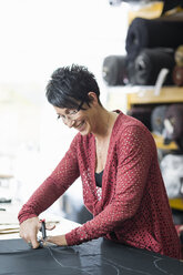 Mature seamstress using scissors to cut textile at work table - CUF38979