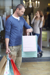 Young woman shopping, man outside holding bags, looking at watch - CUF39008