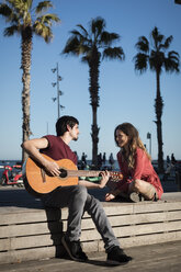 Spanien, Barcelona, lächelndes Paar mit einer Gitarre auf einer Bank an der Strandpromenade sitzend - MAUF01444