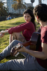 Spanien, Barcelona, junge Frau sitzt auf einer Wiese und beobachtet ihren Freund beim Gitarrenspiel - MAUF01469