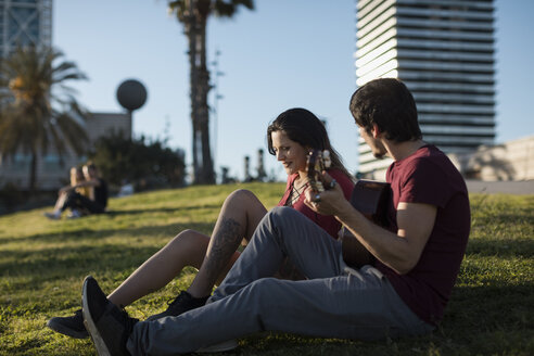 Spanien, Barcelona, junger Mann spielt Gitarre für seine Freundin auf einer Wiese sitzend - MAUF01470