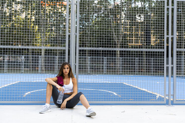 Porträt einer sportlichen jungen Frau, die auf einem Sportplatz sitzt und eine Wasserflasche hält - KKAF01150