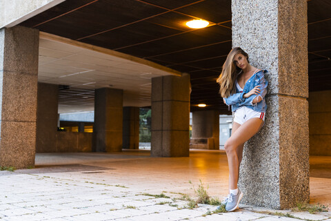 Attraktive junge Frau in Sportkleidung und Jeansjacke lehnt an einer Säule, lizenzfreies Stockfoto