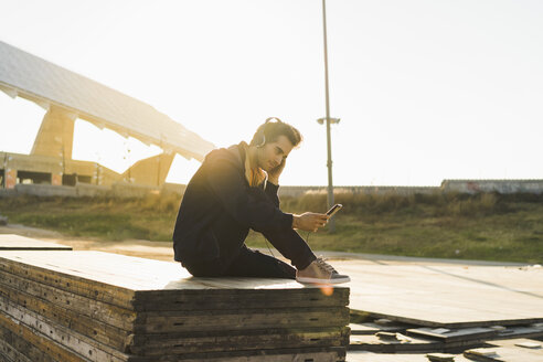 Mann mit Kopfhörern, der auf einem Holzstapel sitzt und ein Smartphone benutzt - AFVF00683