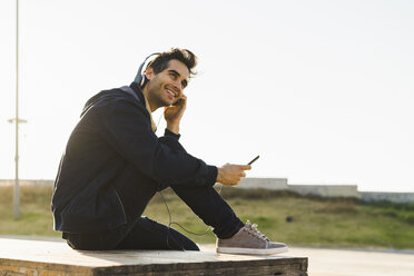 Mann mit Kopfhörern, der auf einem Holzstapel sitzt und ein Smartphone benutzt - AFVF00684