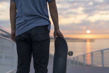 Junger Mann mit Skateboard am Strand bei Sonnenaufgang, Rückansicht - AFVF00718