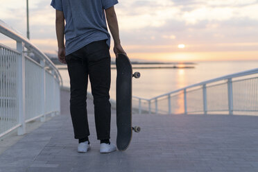 Young man with skateboard at the beach at sunrise, rear view - AFVF00719