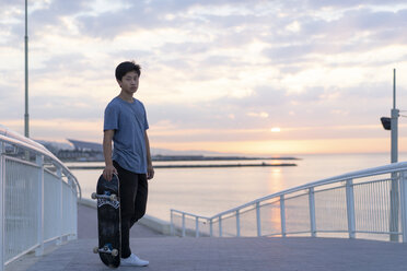 Junger chinesischer Mann mit Skateboard am Strand bei Sonnenaufgang - AFVF00720