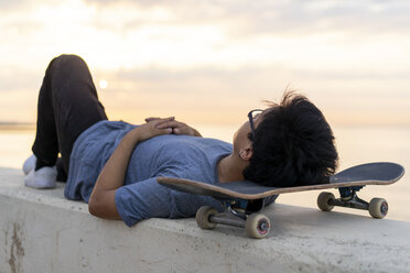 Junger chinesischer Mann mit Skateboard an der Wand liegend am Strand - AFVF00724