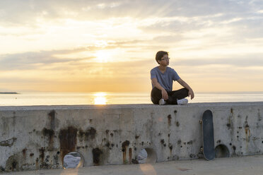 Junger chinesischer Mann sitzt bei Sonnenaufgang auf einer Mauer - AFVF00730
