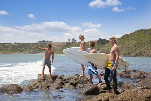 Vier junge männliche Surferfreunde, die von Felsen aus auf das Meer zeigen - ISF16730
