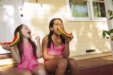 Zwei lachende Mädchen sitzen auf der Veranda eines Hauses mit Wassermelonenscheiben - ISF16763