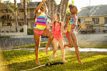 Three girls running and jumping in garden sprinkler - ISF16789