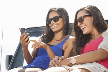 Zwei junge Frauen sitzen auf der Motorhaube eines Jeeps und schauen auf ihr Smartphone - ISF16940
