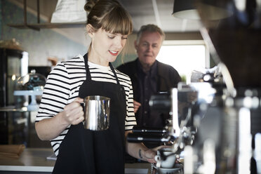 Weibliche Barista hält eine Kanne in der Hand, während sie Kaffee zubereitet, während ein Kunde im Hintergrund im Café steht - MASF08030