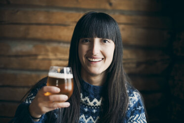 Portrait of smiling woman holding drinking beer glass while sitting at log cabin - MASF08128