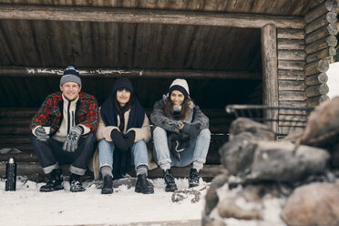 Full length portrait of friends sitting in log cabin against trees during winter - MASF08134