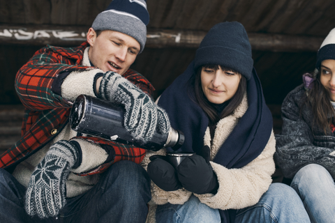 Mann serviert weiblichen Freunden ein Getränk, während er in einer Blockhütte sitzt, lizenzfreies Stockfoto