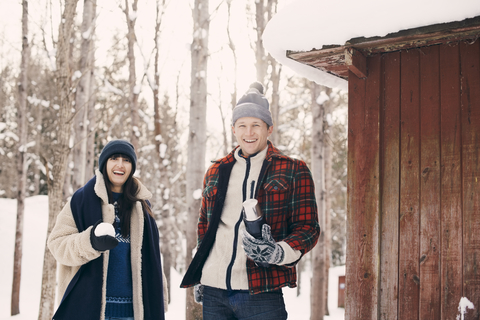 Porträt von glücklichen Freunden, die einen Schneeball halten, während sie im Winter vor einer Blockhütte stehen, lizenzfreies Stockfoto