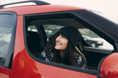 Lächelnde Frau, die wegschaut, während sie in einem roten Auto sitzt, lizenzfreies Stockfoto