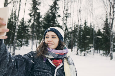 Smiling woman taking selfie through mobile phone while standing against trees on snowy field - MASF08179