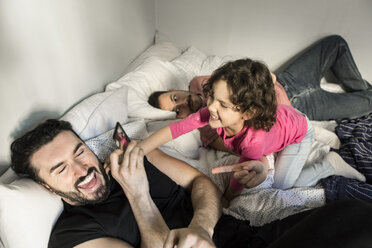 Daughter lying by man playing with father holding smart phone on bed in bedroom - MASF08206