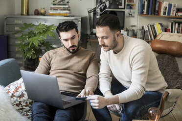 Gay couple shopping online with credit card through laptop while relaxing on chairs at home - MASF08209