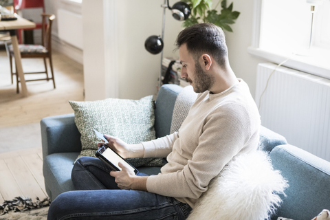 Junger Mann beim Online-Einkauf auf einem digitalen Tablet mit Kreditkarte, während er zu Hause auf dem Sofa sitzt, lizenzfreies Stockfoto