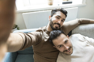 Lächelndes homosexuelles Paar, das ein Selfie macht, während es sich auf dem Sofa im Wohnzimmer zu Hause entspannt - MASF08216