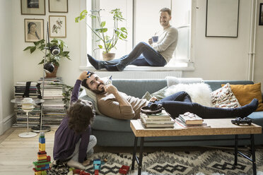 Girl playing with fathers while man sitting on window sill in living room - MASF08221